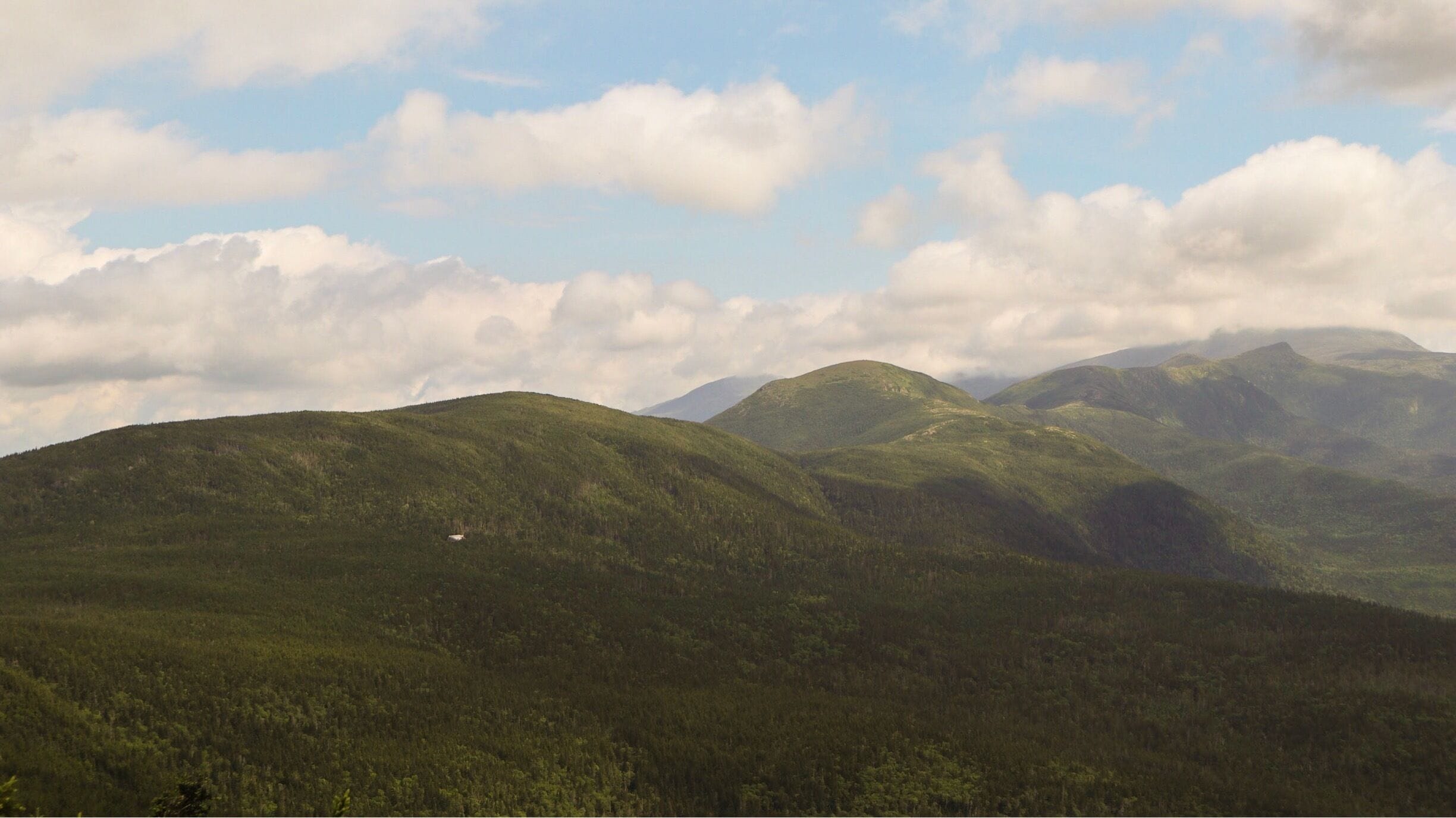 A mountainous landscape with dense forests and rolling hills under a partly cloudy sky.