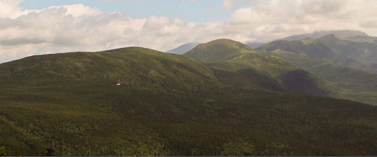 A mountainous landscape with dense forests and rolling hills under a partly cloudy sky.