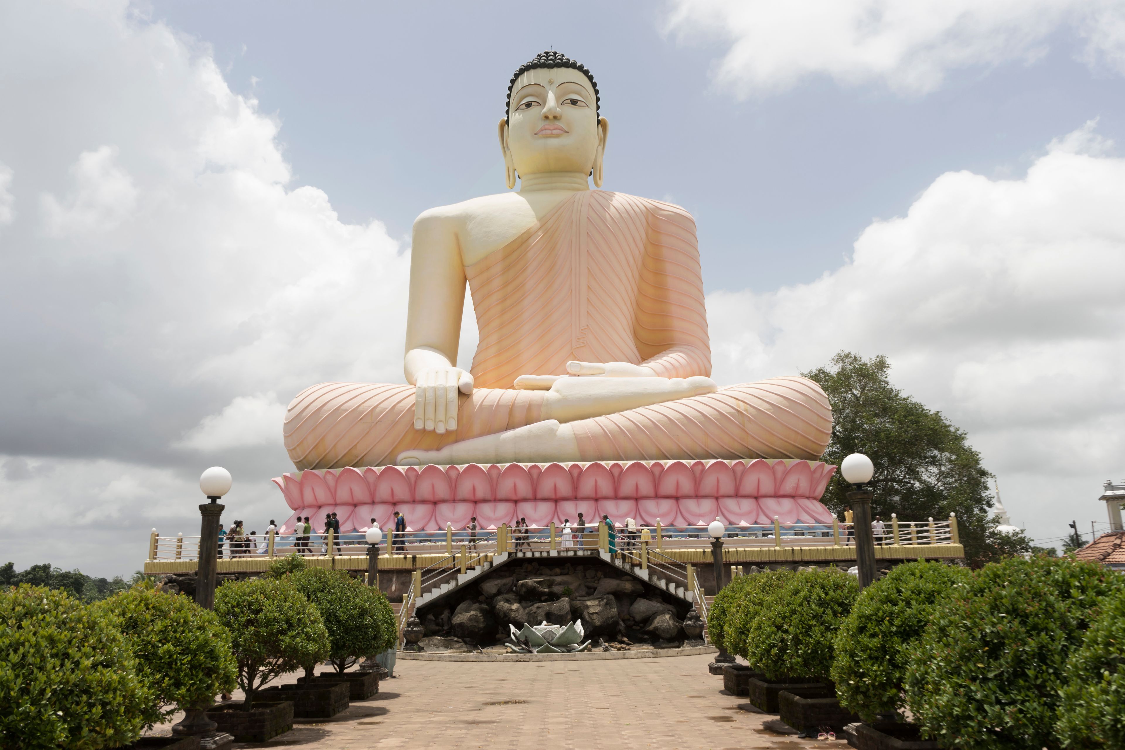 A large seated Buddha statue with a lotus base, surrounded by a paved walkway and small trees.