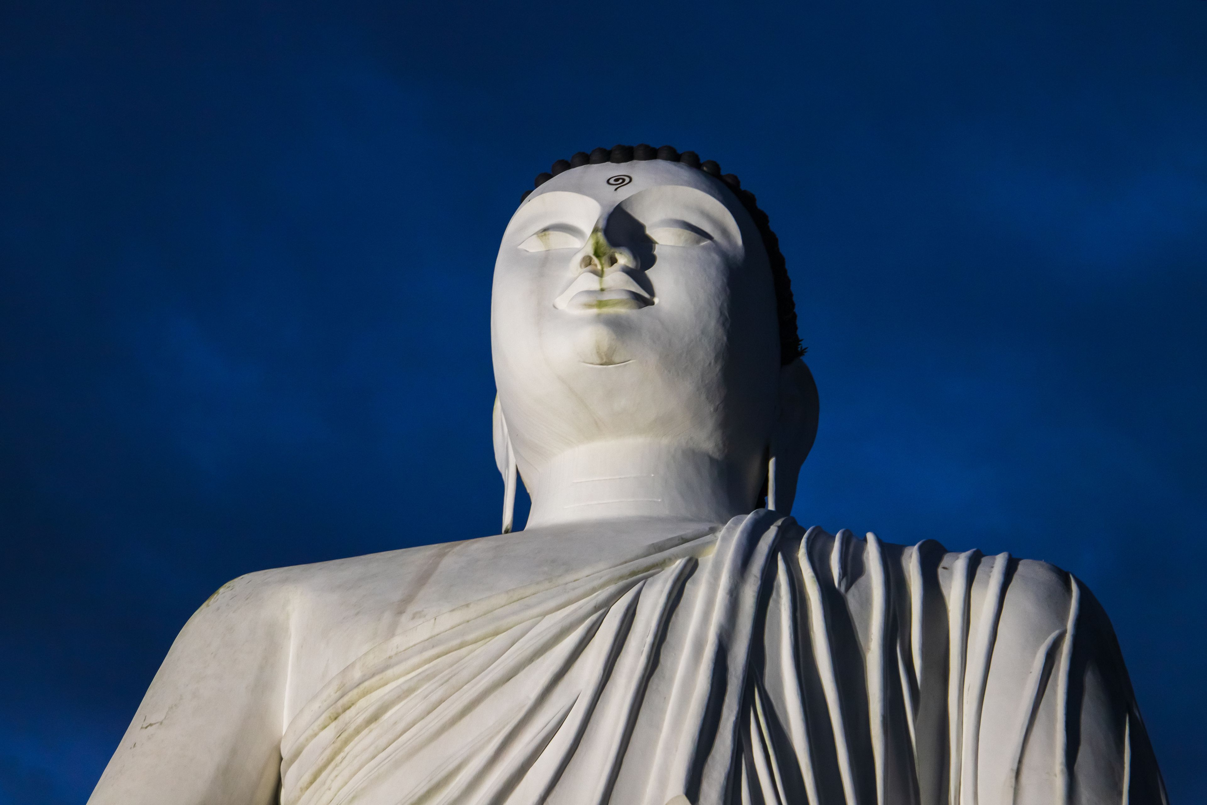 A large white statue with a serene expression, draped in a white cloth, set against a clear blue sky.