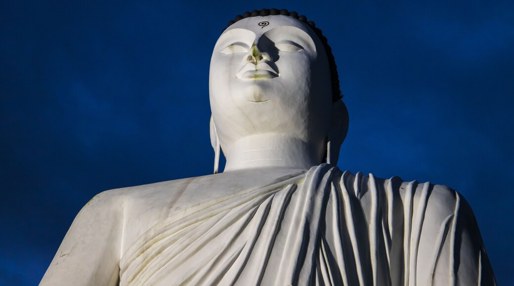 A large white statue with a serene expression, draped in a white cloth, set against a clear blue sky.