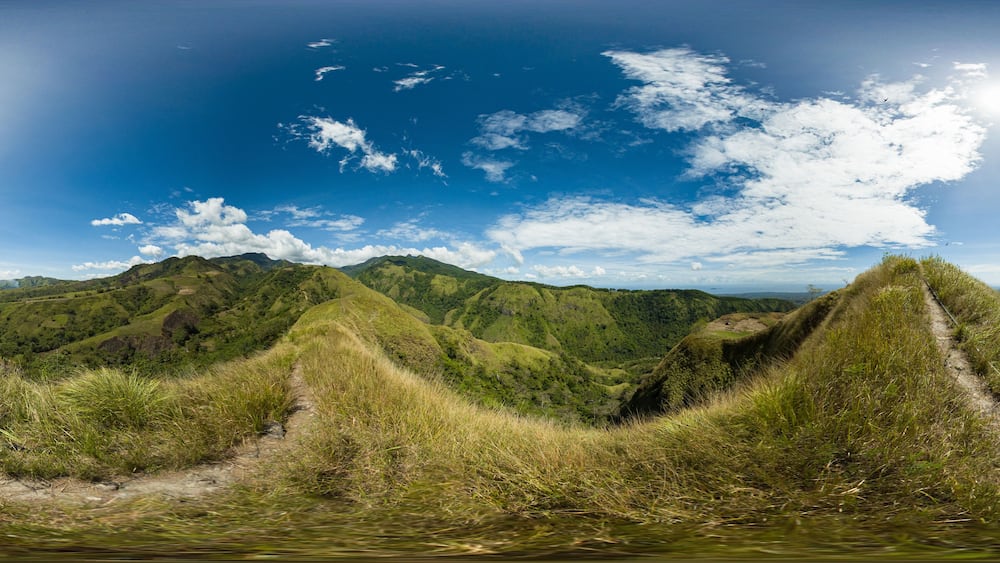 Une vue panoramique sur des collines verdoyantes, avec un ciel bleu dégagé et quelques nuages épars.