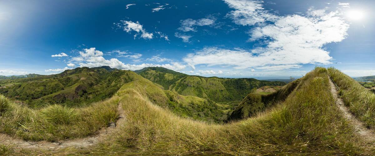 A panoramic view of lush green hills with a clear blue sky and scattered clouds.