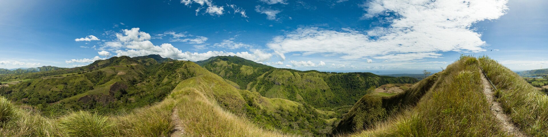 A panoramic view of lush green hills with a clear blue sky and scattered clouds.