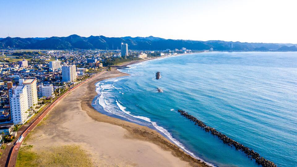 A coastal city with a beach, a rocky breakwater, and mountains in the background.