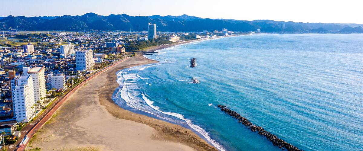 A coastal city with a beach, a rocky breakwater, and mountains in the background.