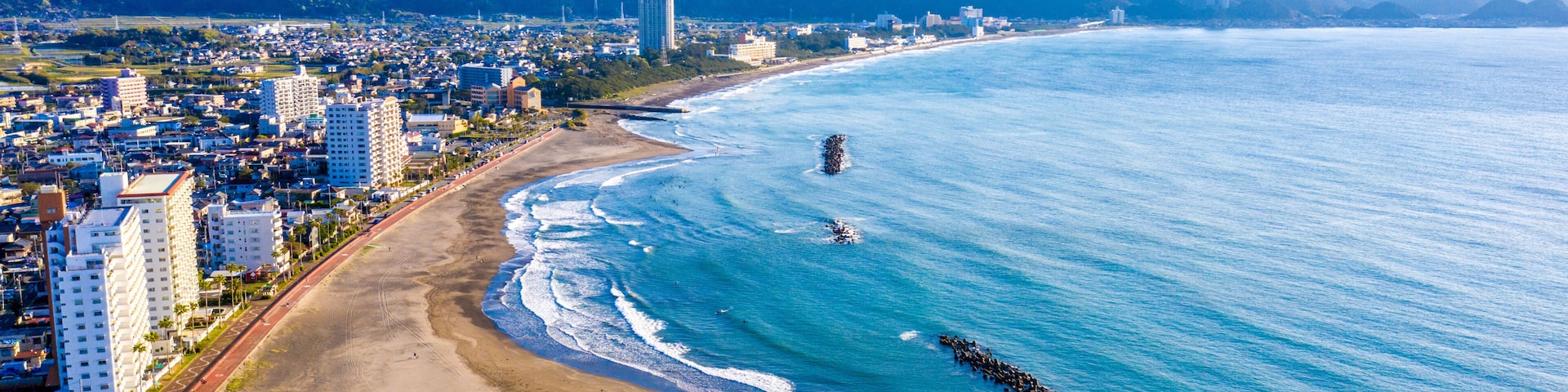 A coastal city with a beach, a rocky breakwater, and mountains in the background.