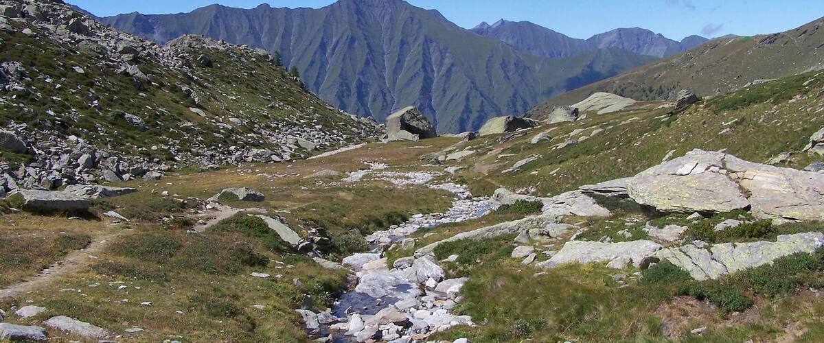 A rocky mountain path with a stream, surrounded by green grass and mountains in the background.