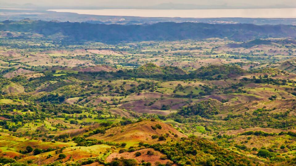 A vast landscape with rolling hills, patches of greenery, and distant mountains under a cloudy sky.