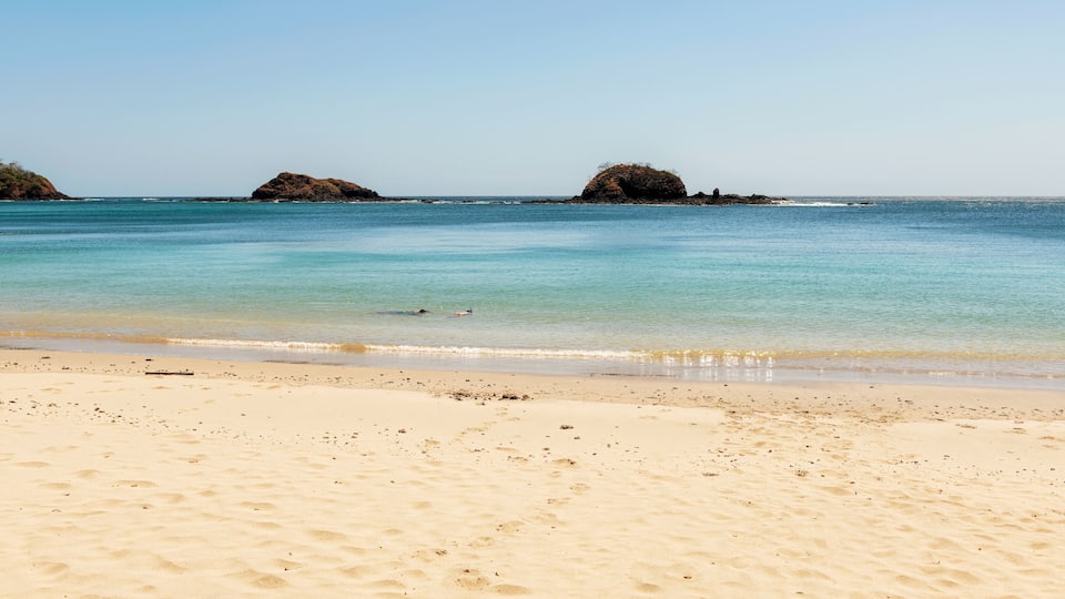 A sandy beach with clear turquoise water, two rocky outcrops, and a clear sky.