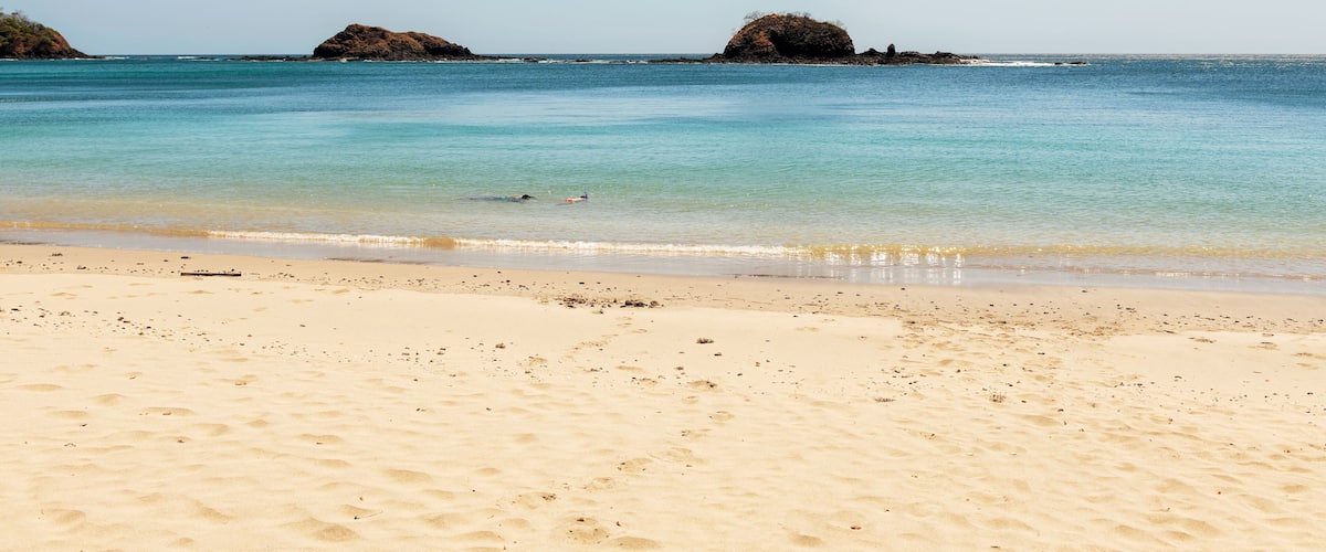 A sandy beach with clear turquoise water, two rocky outcrops, and a clear sky.