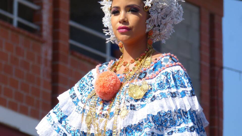 A person in traditional attire with a white and blue patterned dress, adorned with gold jewelry and a decorative headpiece.