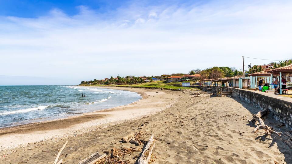 A sandy beach with waves, a wooden walkway, and houses in the background.