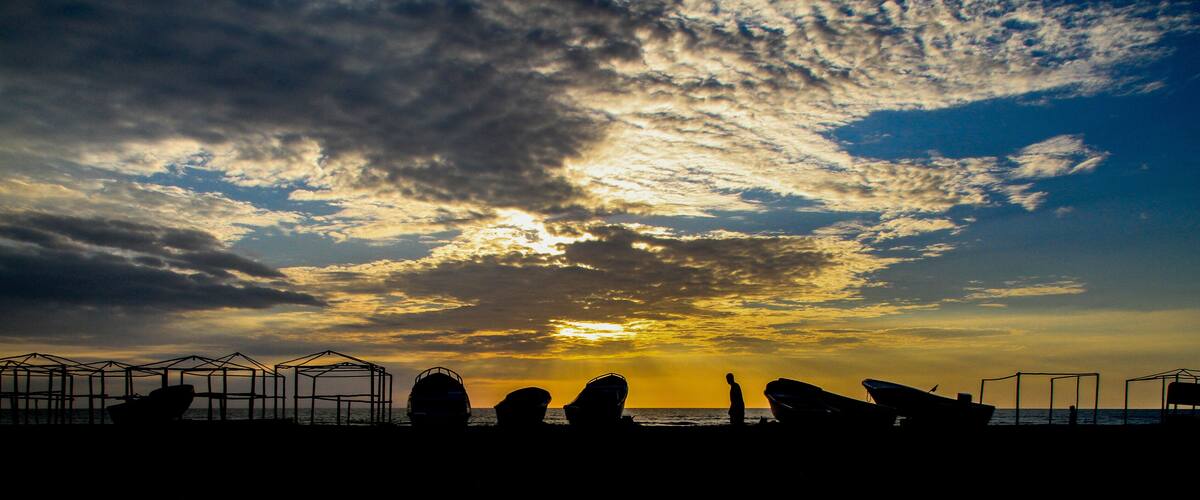 A sunset over a beach with boats and a person silhouetted against the sky.