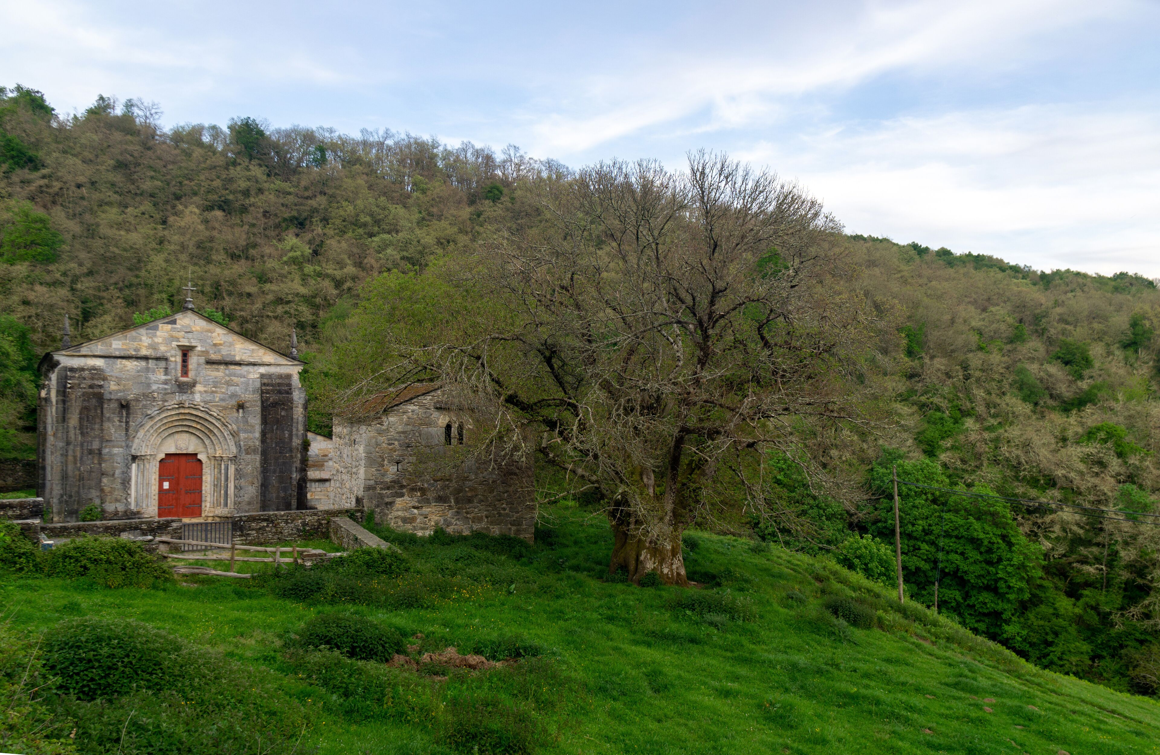A stone church with a red door, surrounded by lush green grass and trees on a hillside.