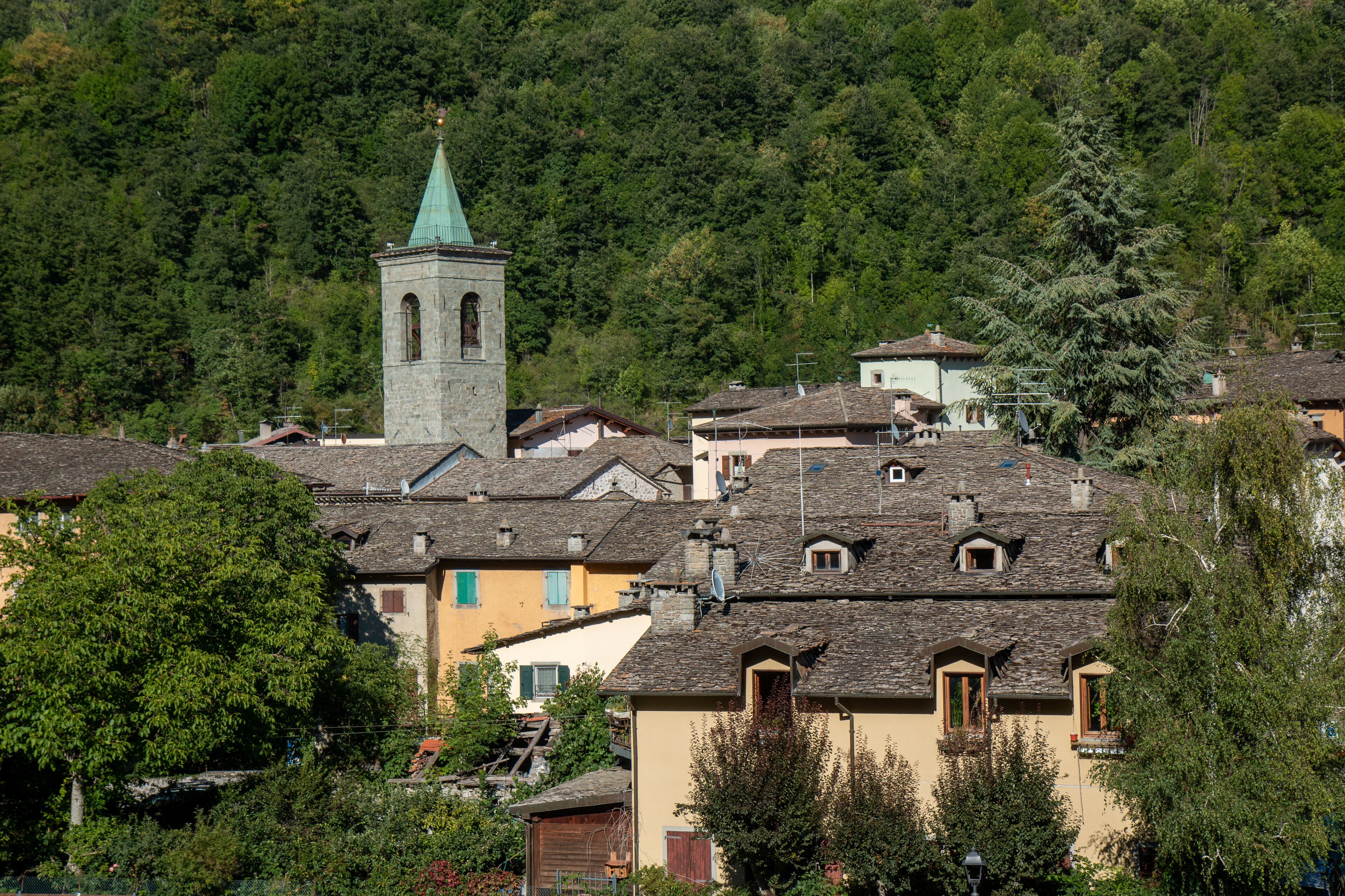 A village with stone buildings, a church with a green spire, and a forested hillside.
