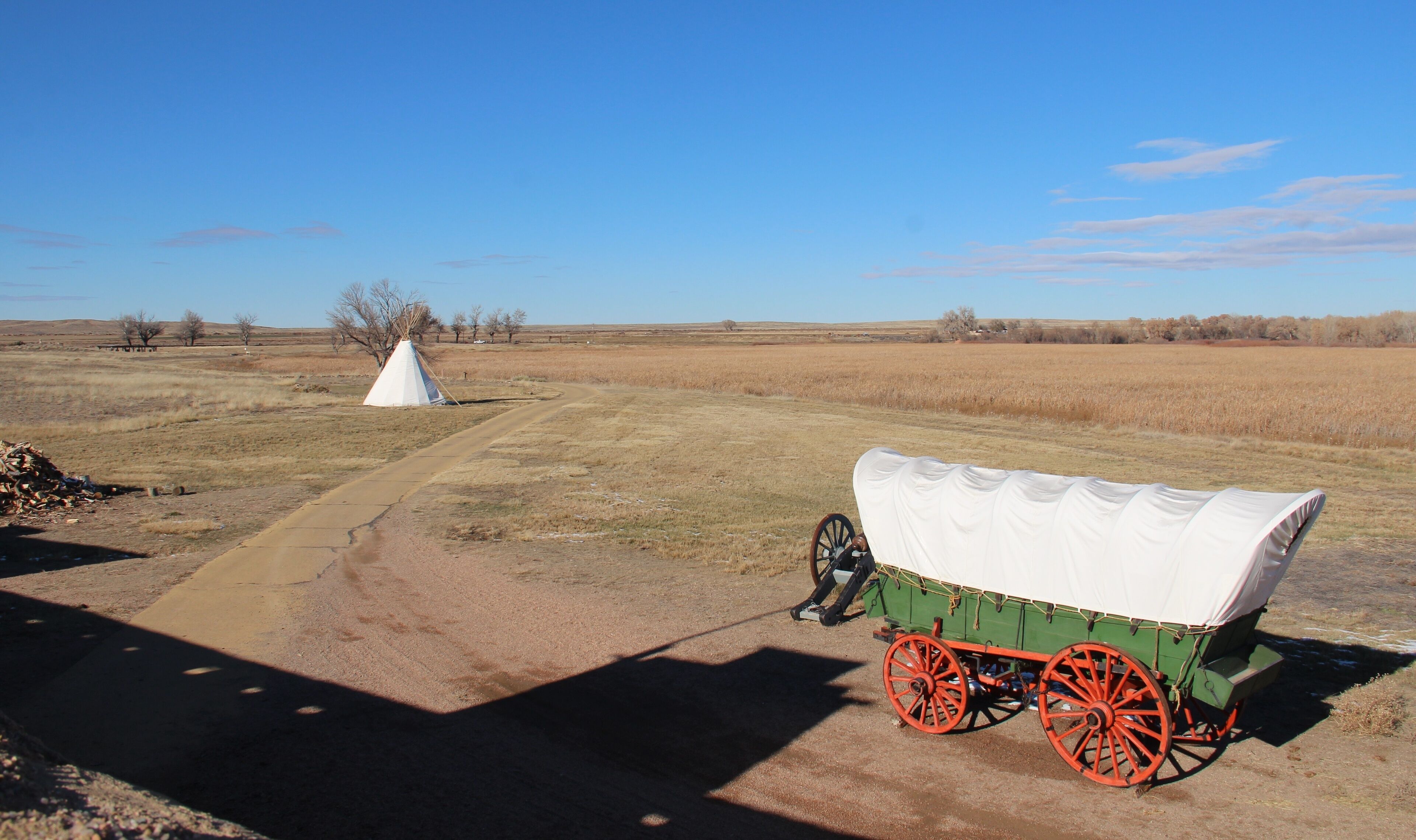 A covered wagon with red wheels parked on a dirt road in a rural field with a white tipi in the background.