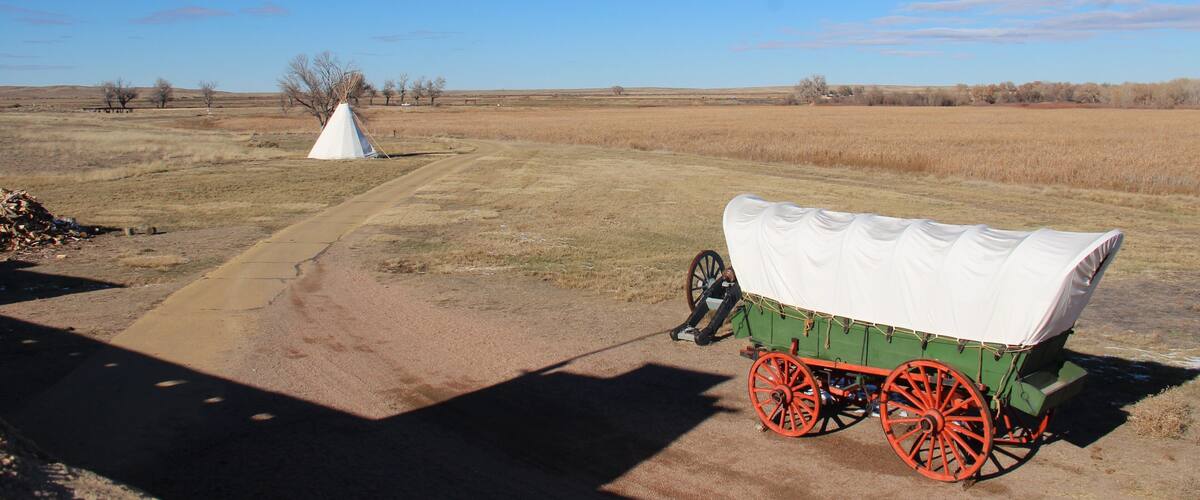 A covered wagon with red wheels parked on a dirt road in a rural field with a white tipi in the background.
