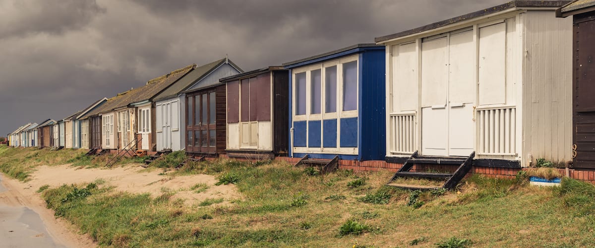 A row of beach huts with a sandy beach and grassy area in the foreground.