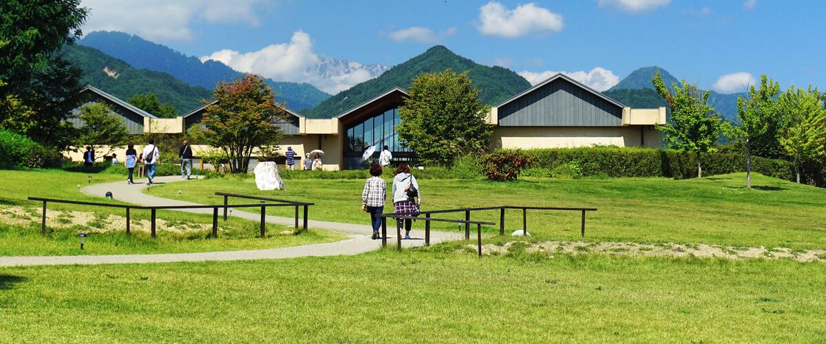 A grassy field with a pathway, buildings, and mountains in the background.