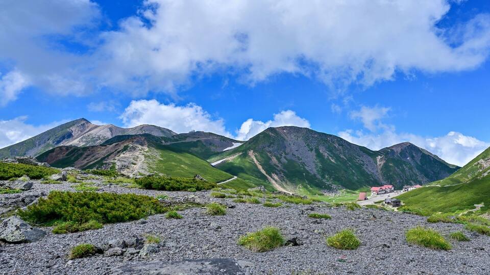 A mountainous landscape with rocky terrain, green vegetation, and a small building with a red roof.