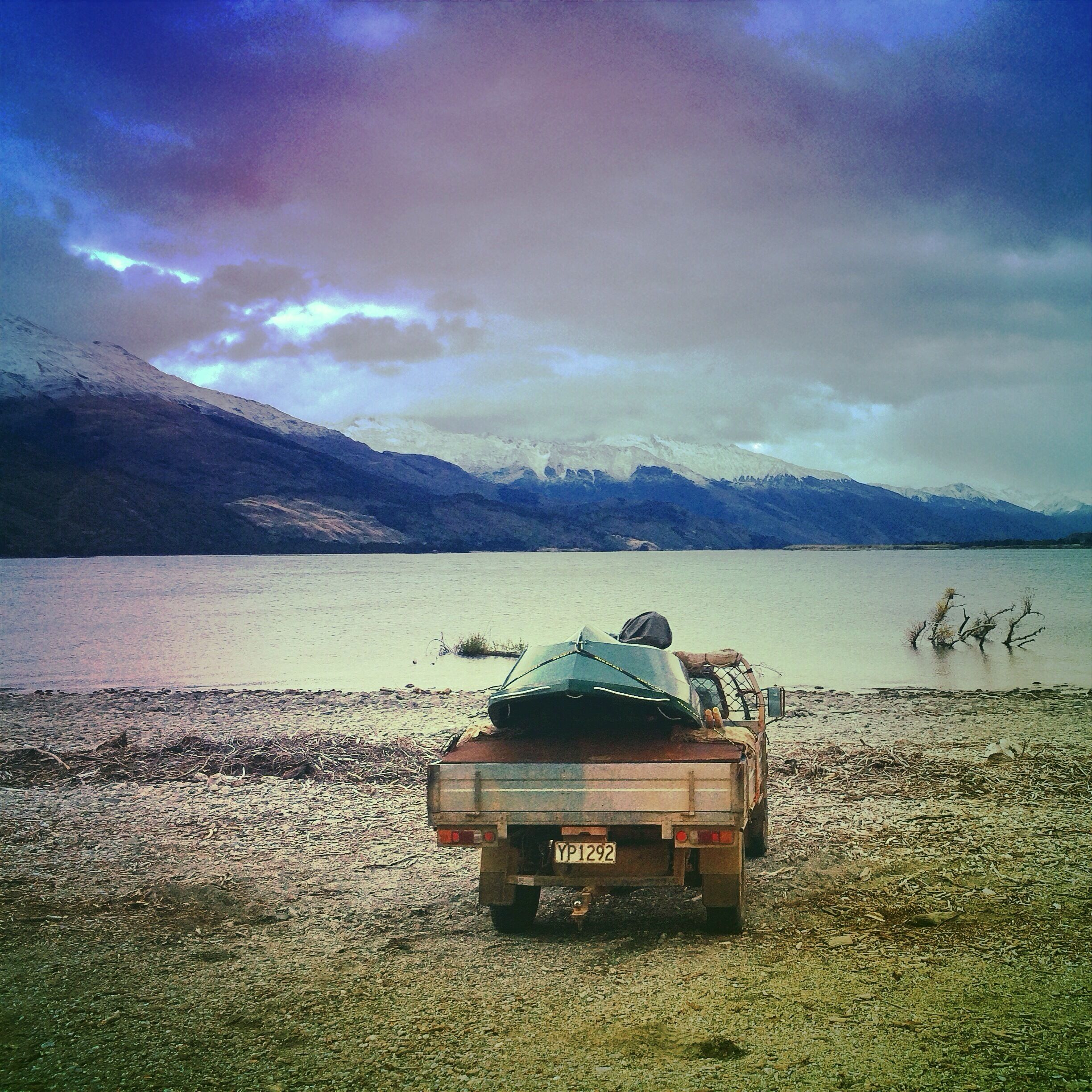 A truck parked on a gravel beach with a mountainous backdrop and a body of water.