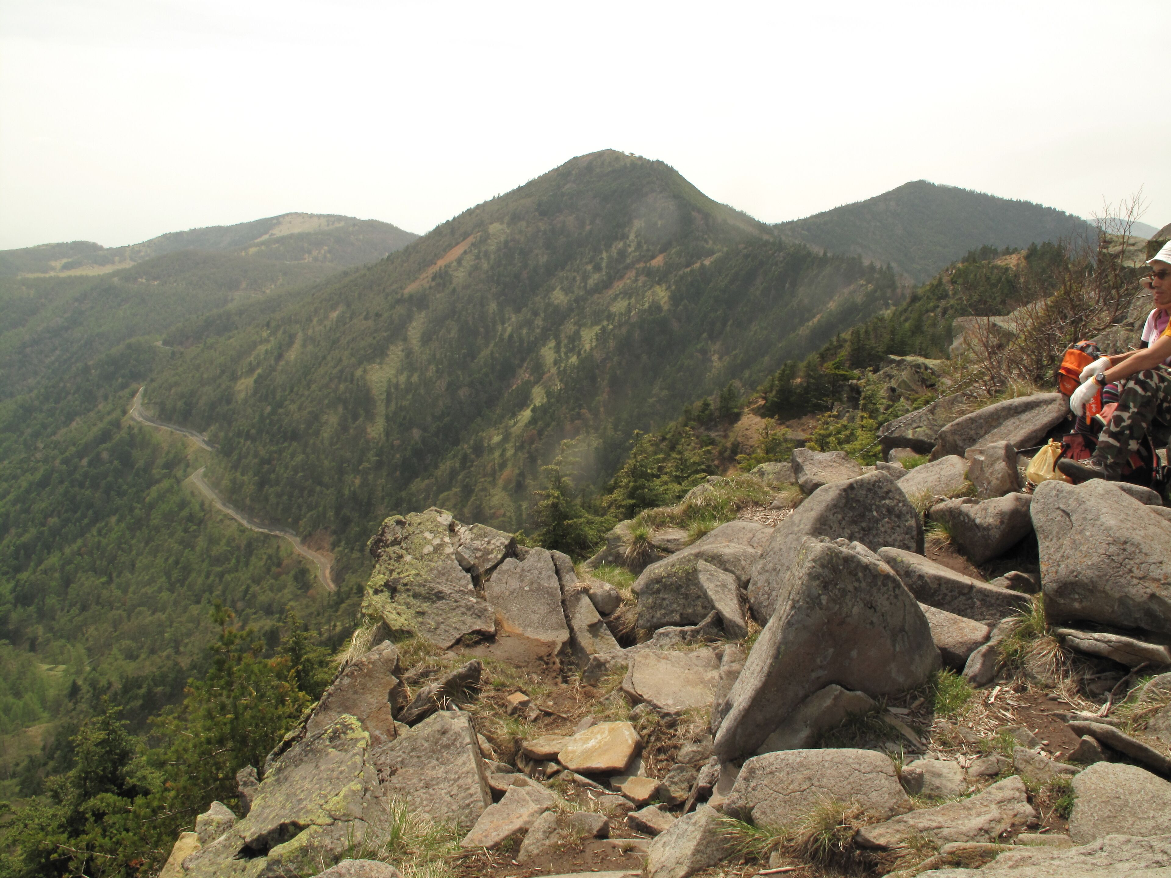 A hiker on a rocky mountain path with a view of a valley and forested hills in the background.