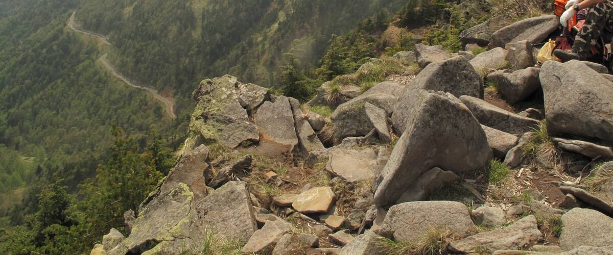 A hiker on a rocky mountain path with a view of a valley and forested hills in the background.