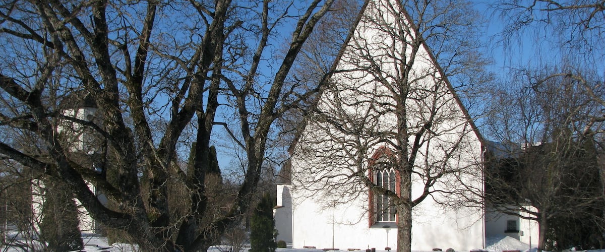 A snow-covered cemetery with a prominent church featuring a pointed steeple and a large tree in the foreground.