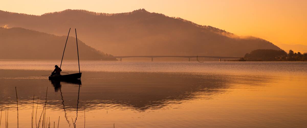 A calm lake at sunset with a bridge in the distance and hills in the background.