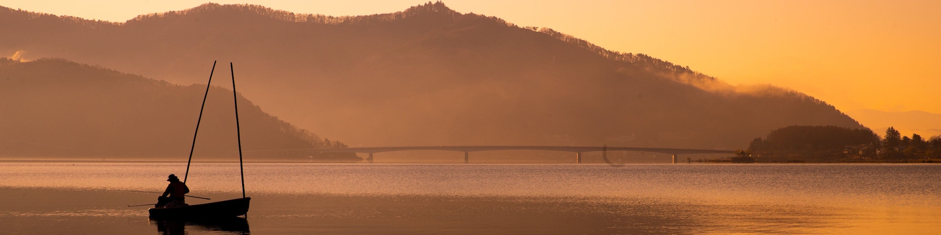 A calm lake at sunset with a bridge in the distance and hills in the background.