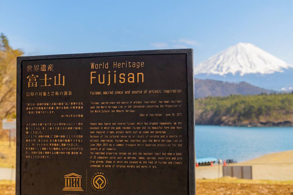 A signboard with information about Fujisan, a World Heritage site, in front of a scenic view of a lake and a snow-capped mountain.