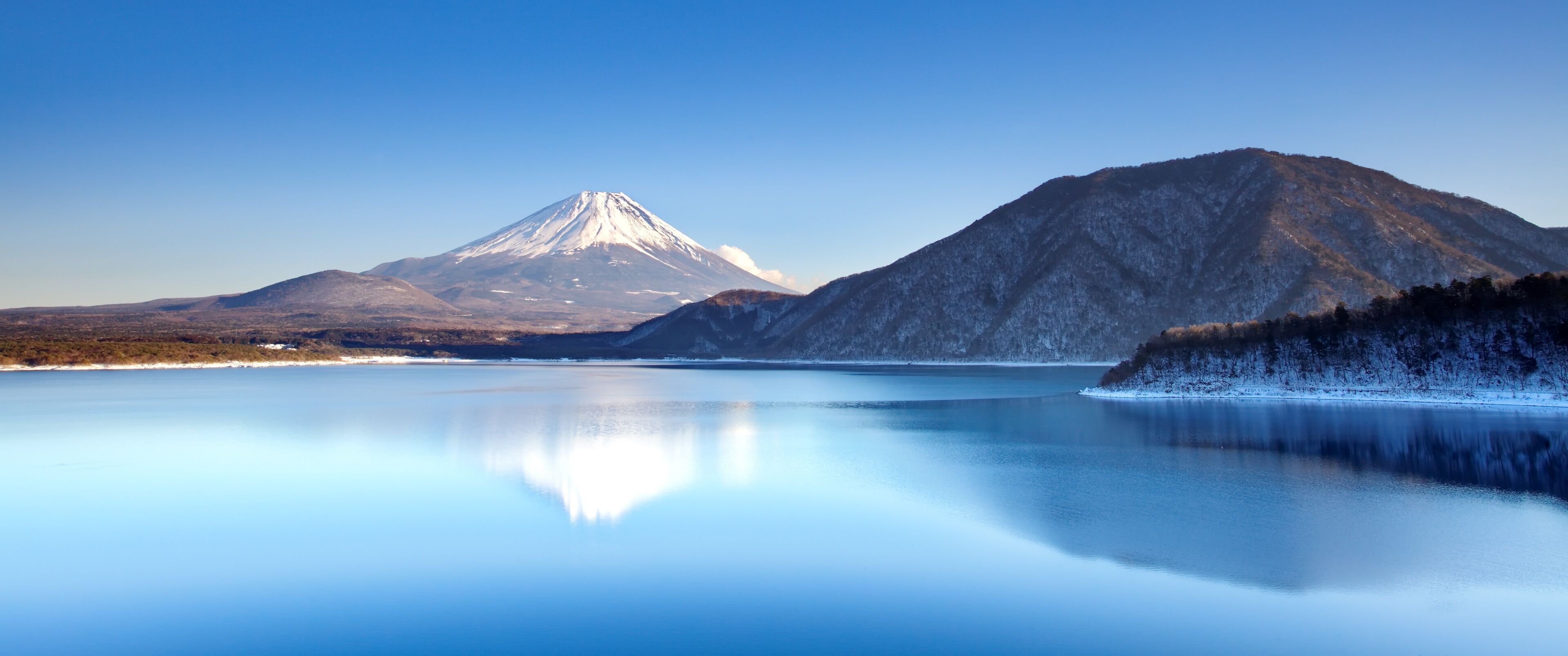 A calm lake reflecting a snow-capped mountain and a forested hill.