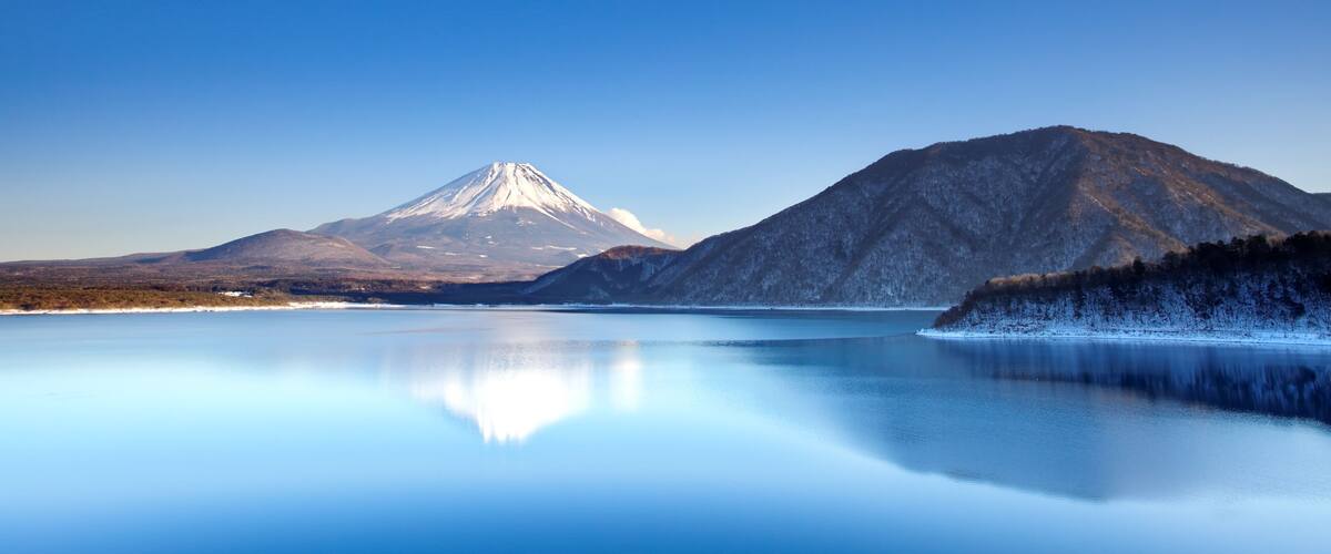 A calm lake reflecting a snow-capped mountain and a forested hill.