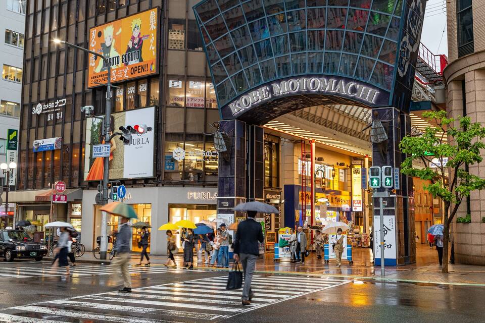A busy urban street with a prominent Kobe Momochi entrance, various storefronts, and pedestrians crossing the street.