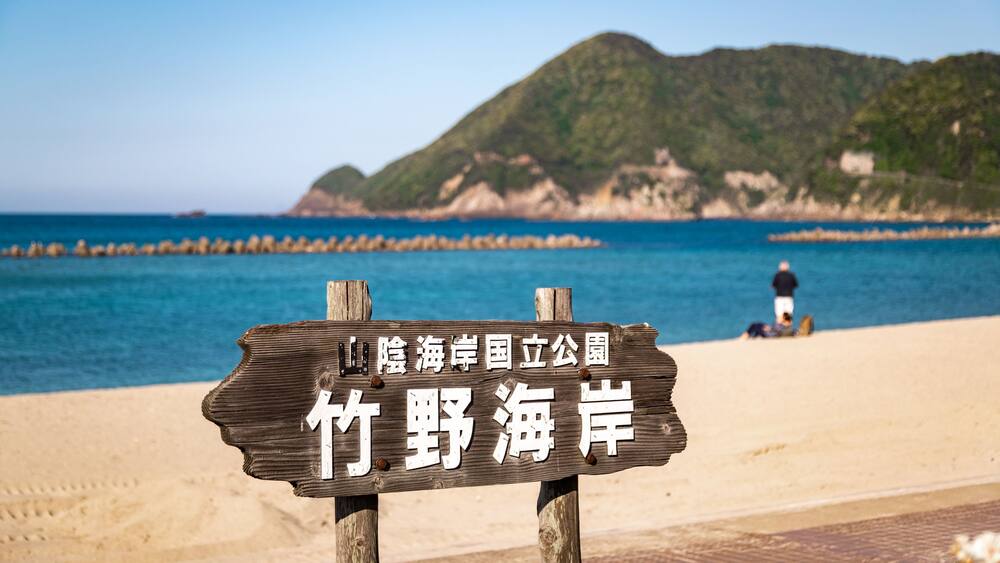 Un panneau en bois orné de caractères chinois, sur une plage, avec une montagne en arrière-plan et la mer.