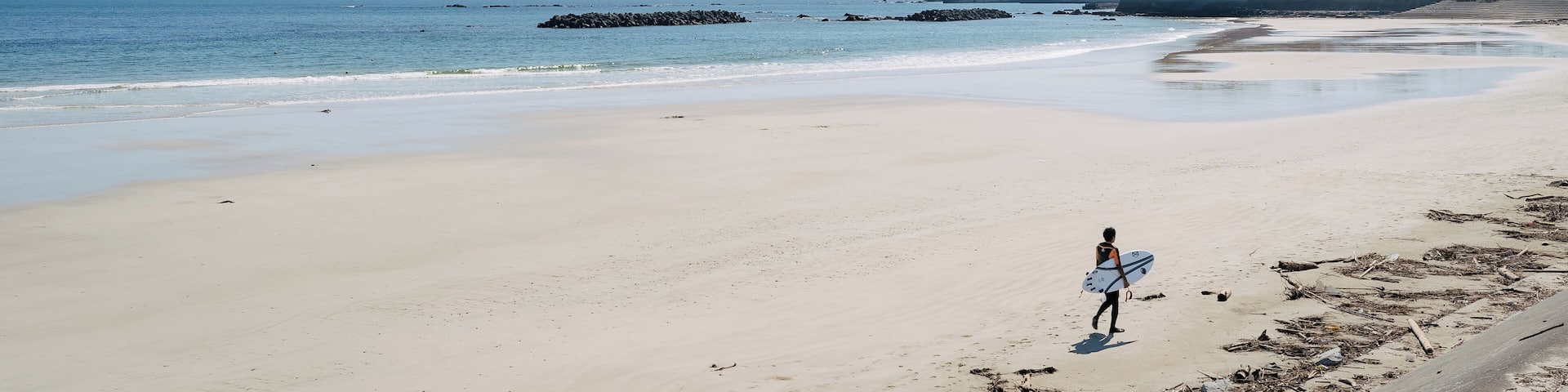 A beach with a person carrying a surfboard, calm water, and a distant rocky outcrop.