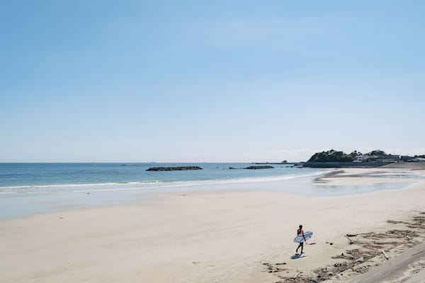 Ein Strand mit einer Person, die einen Surfbrett trägt, ruhigem Wasser und einer fernen Felsenformation.