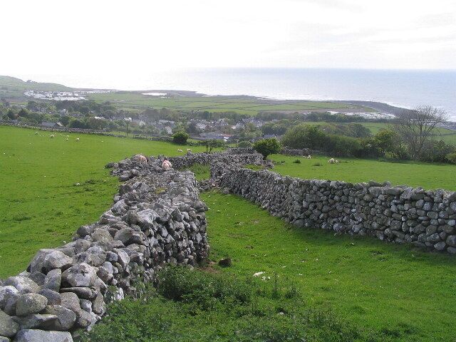 Un mur de pierres dans un champ verdoyant, avec une vue sur la côte en arrière-plan.