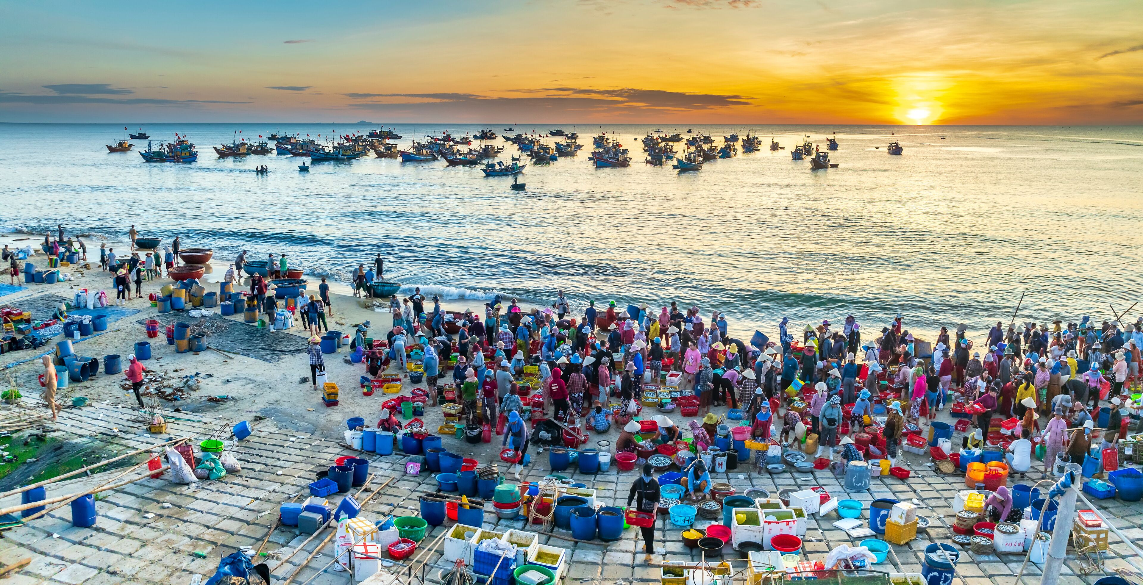 A bustling coastal fishing village at sunset with numerous boats and people engaged in fishing activities.