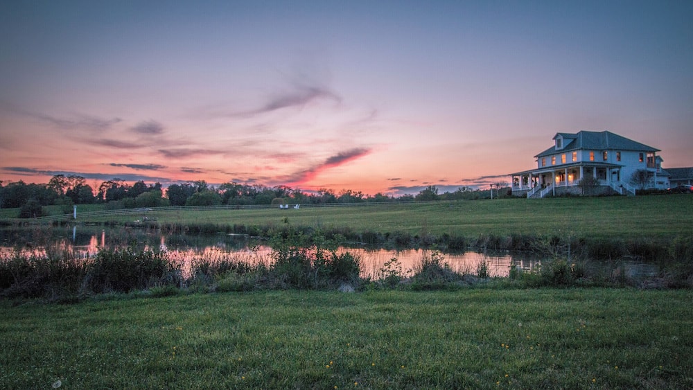A large house with a porch, a pond, and a sunset sky.