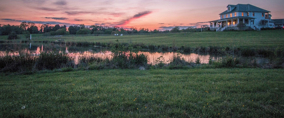 A large house with a porch, a pond, and a sunset sky.