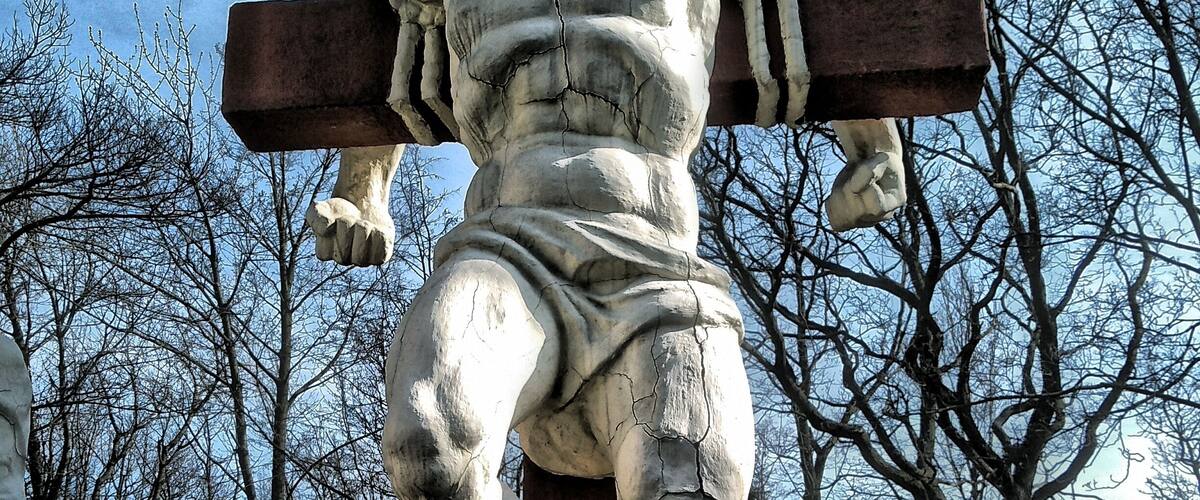 A statue of a muscular figure carrying a cross, set against a backdrop of leafless trees and a clear sky.