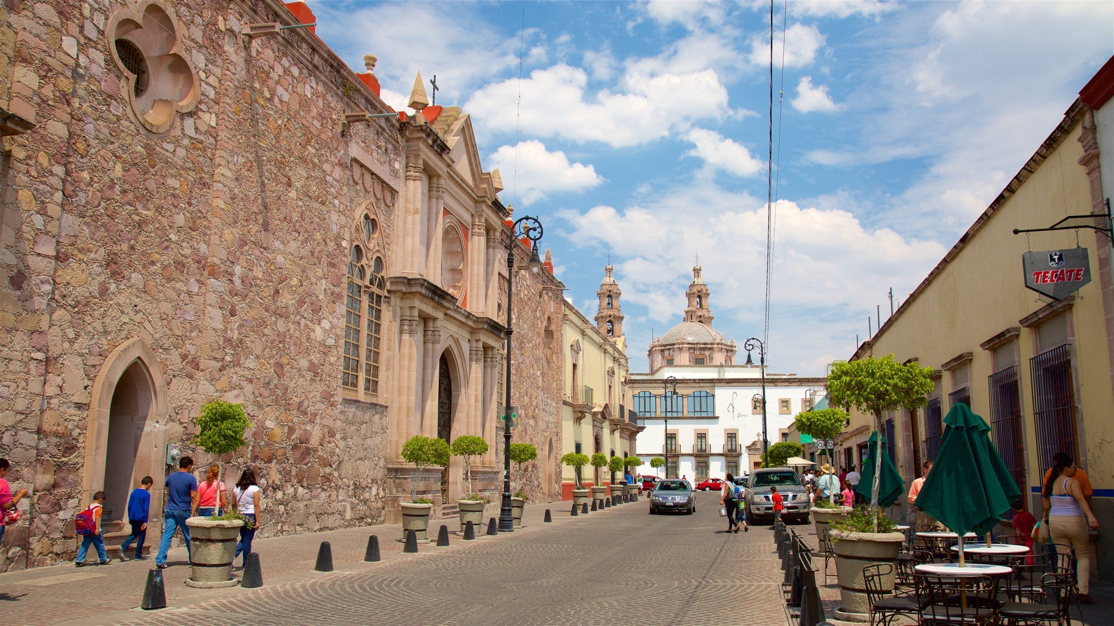 A cobblestone street lined with historic stone buildings, a prominent church with a dome, and people walking along the sidewalk.