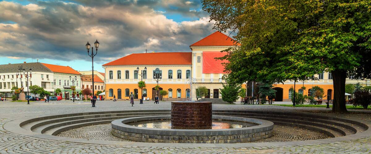 A cobblestone square with a central fountain, surrounded by historic buildings with red roofs.