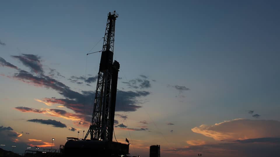 A large industrial drilling rig silhouetted against a sunset sky with scattered clouds.