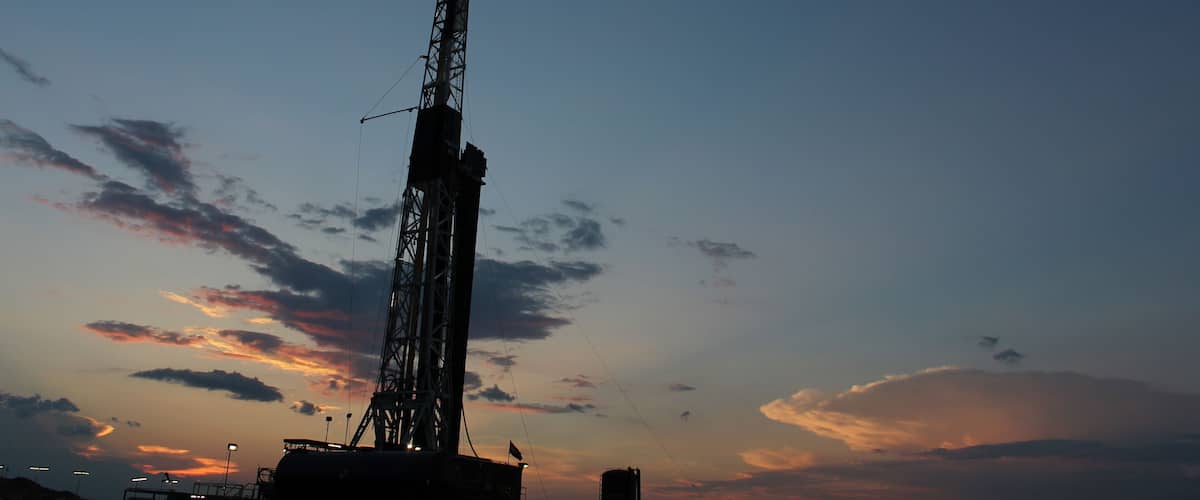 A large industrial drilling rig silhouetted against a sunset sky with scattered clouds.