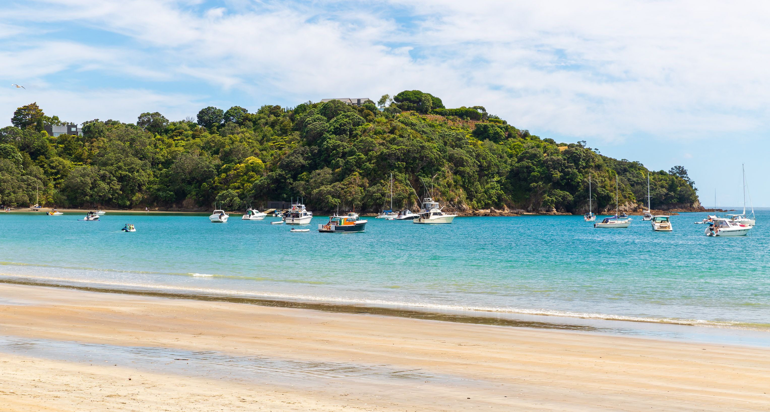 A beach with boats on calm turquoise waters, a sandy shore, and a forested hill in the background.