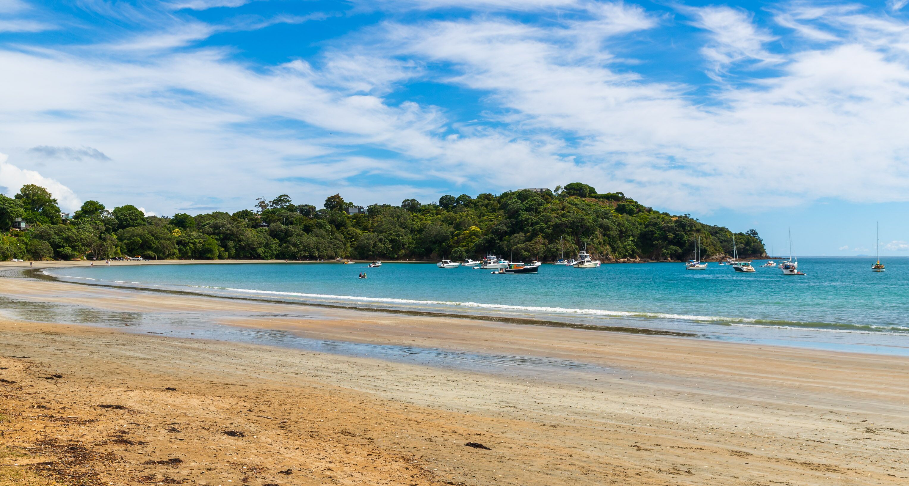 Uma praia arenosa com barcos na água e uma colina arborizada ao fundo.