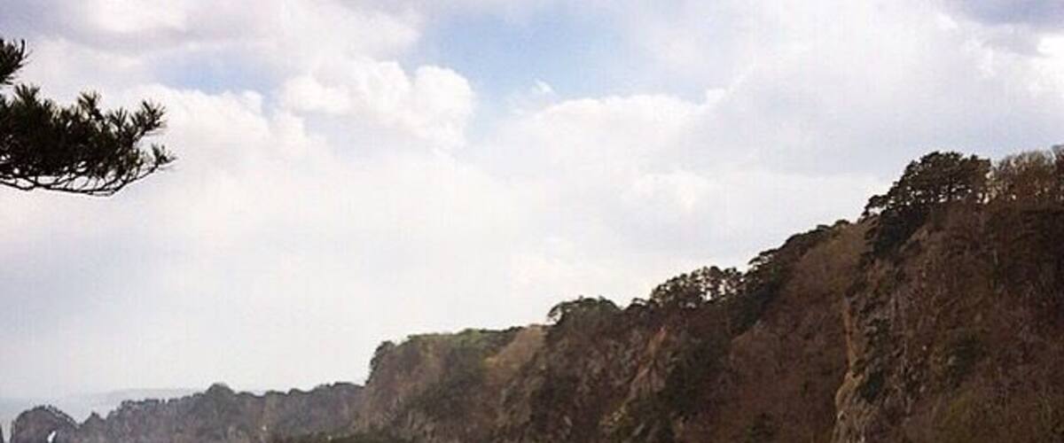 A coastal landscape with rugged cliffs, dense greenery, and a calm sea.